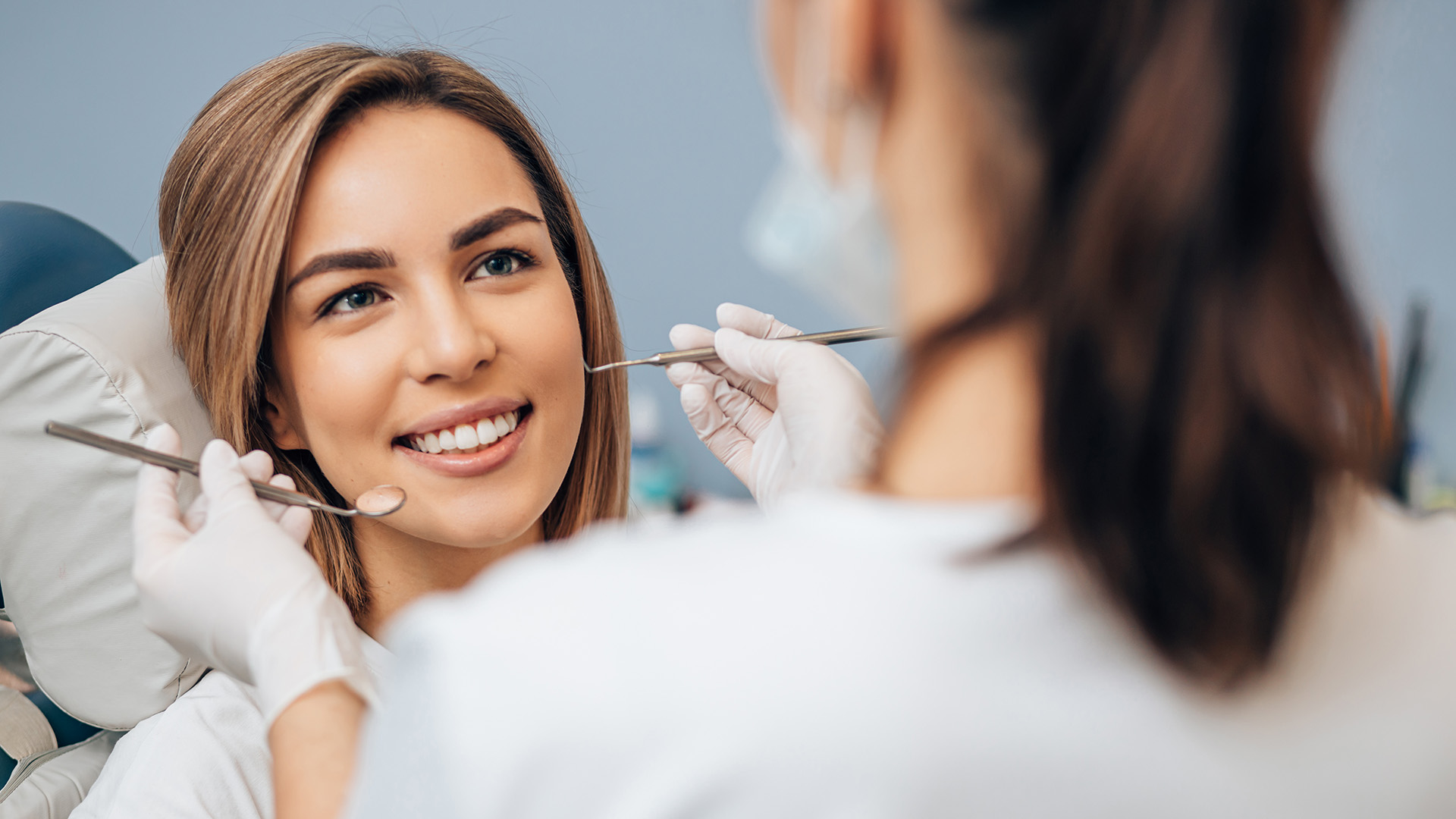 Adolescent Dental Care. A teenager sits in the dentist's chair and looks toward the dentist who is performing an exam.