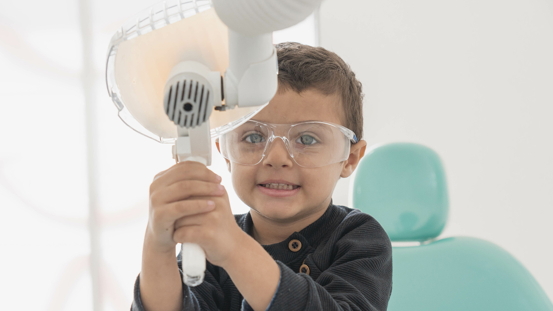 Primera visita? Que esperar. A smiling child holds onto the exam light while wearing clear protective glasses.