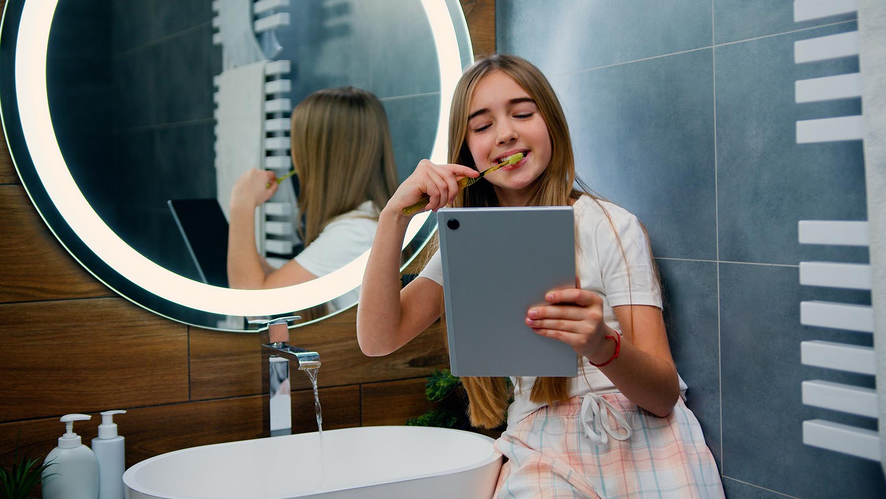 Reading Our Blog and Keeping Your Teeth Clean. A teenager reads from a tablet while brushing her teeth.